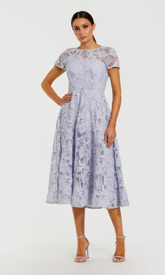 A woman wearing the Midi Party Dress: Mac Duggal 50073 in lilac, featuring floral lace, short sleeves, and an elegant a-line midi skirt, paired with high heels and standing against a plain white background.