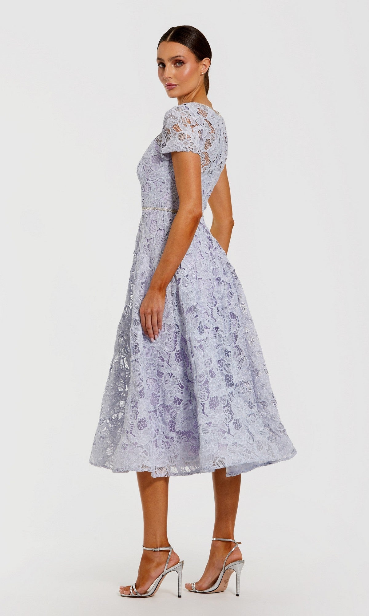 A woman stands in profile against a plain white background, wearing the Midi Party Dress: Mac Duggal 50073—a light purple floral lace midi dress featuring short sleeves and an elegant A-line skirt—paired with silver high-heeled sandals.
