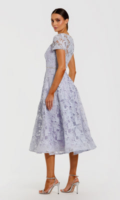 A woman stands in profile against a plain white background, wearing the Midi Party Dress: Mac Duggal 50073—a light purple floral lace midi dress featuring short sleeves and an elegant A-line skirt—paired with silver high-heeled sandals.