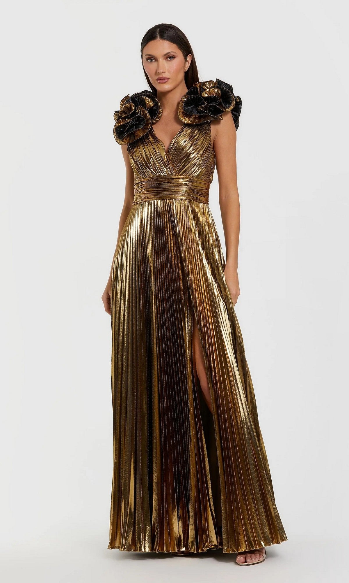 A woman wears the Mac Duggal 50084 long formal dress in metallic gold, featuring pleats, a deep V-neck, bold black floral shoulder accents, and a high slit, standing against a plain light background.