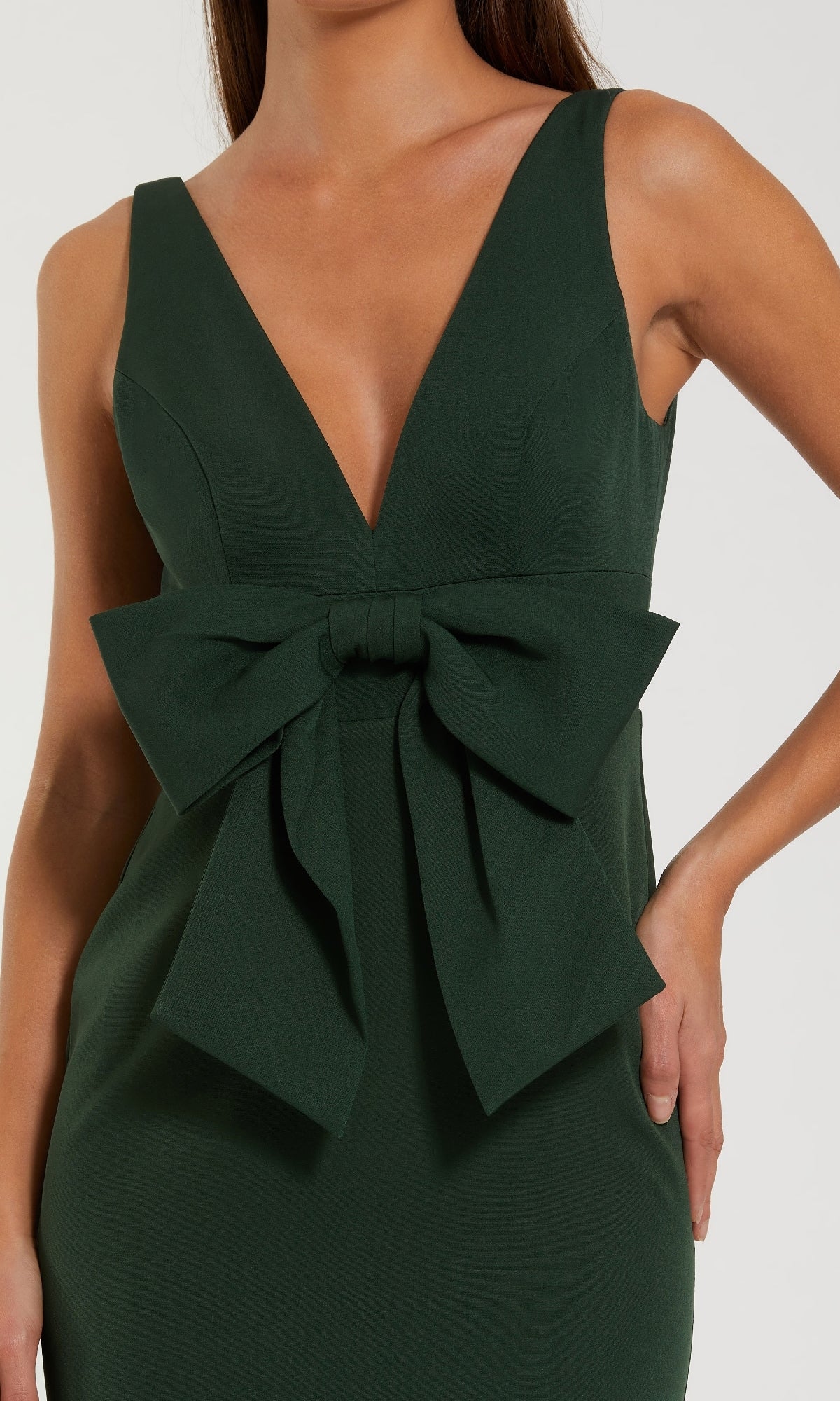 A person wearing the Mac Duggal 50087 Bow-Front Green Formal Dress, featuring an elegant sleeveless design, deep V-neckline, long silhouette, and a striking bow detail at the waist.