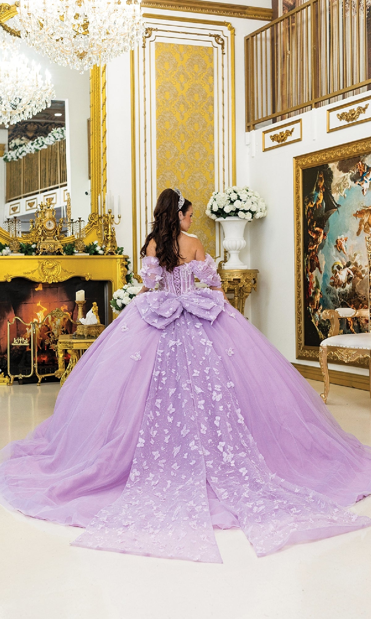 A woman wearing the Purple Quinceanera Dress 5008 by Dancing Queen, featuring butterfly appliques and a large bow, stands in an ornate room with gold accents, a fireplace, mirrors, and classical artwork.