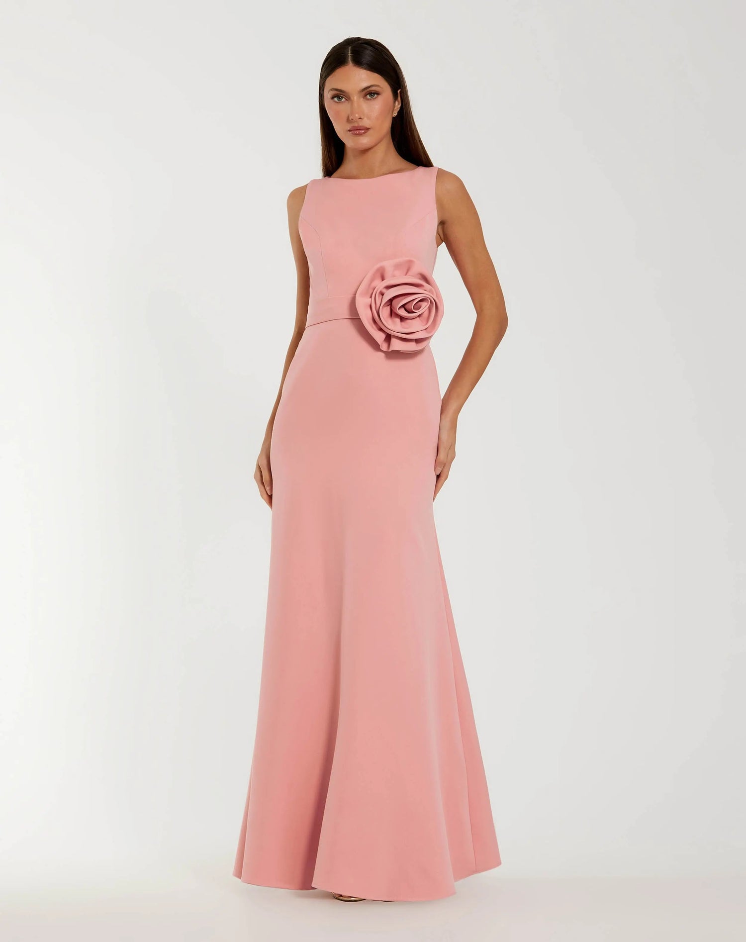 A woman wearing the Mac Duggal 50097 long formal dress in rose—a sleeveless, floor-length crepe gown with a large fabric rose detail at the waist—stands against a plain light background.