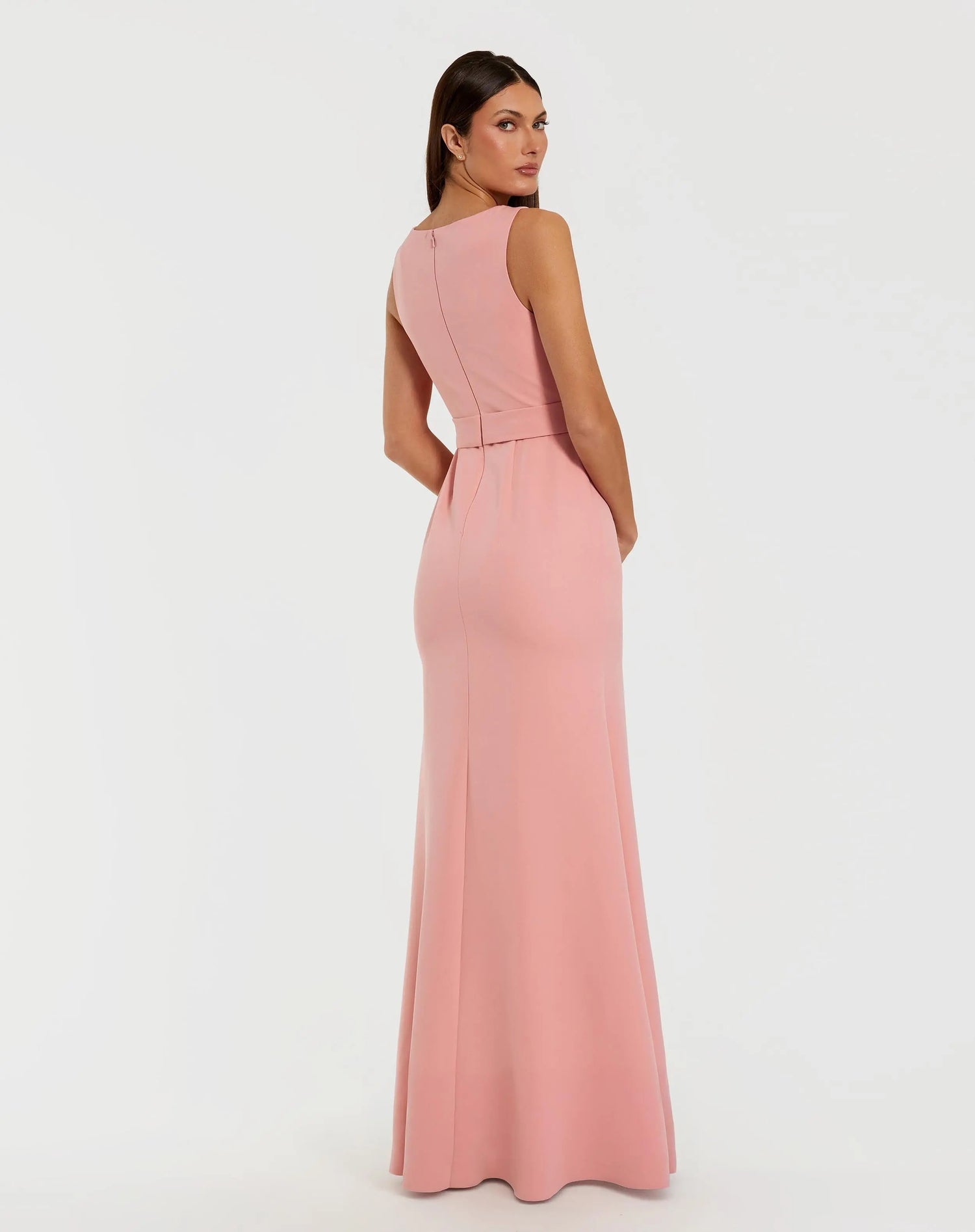 A woman is seen from behind, glancing over her left shoulder while wearing the Mac Duggal 50097 Long Formal Dress—a sleeveless, floor-length crepe gown in a light pink pastel shade—set against a plain white background.