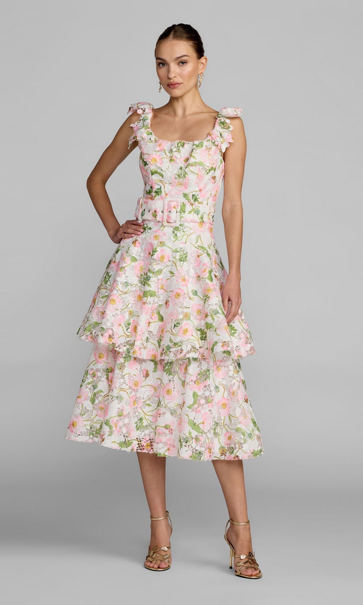 A woman stands in the Mac Duggal 50112 Midi Party Dress in White Multi, featuring a sleeveless, tiered floral lace design with pink, green, and white patterns. She wears gold heels and poses against a plain gray background.