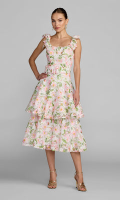 A woman stands in the Mac Duggal 50112 Midi Party Dress in White Multi, featuring a sleeveless, tiered floral lace design with pink, green, and white patterns. She wears gold heels and poses against a plain gray background.