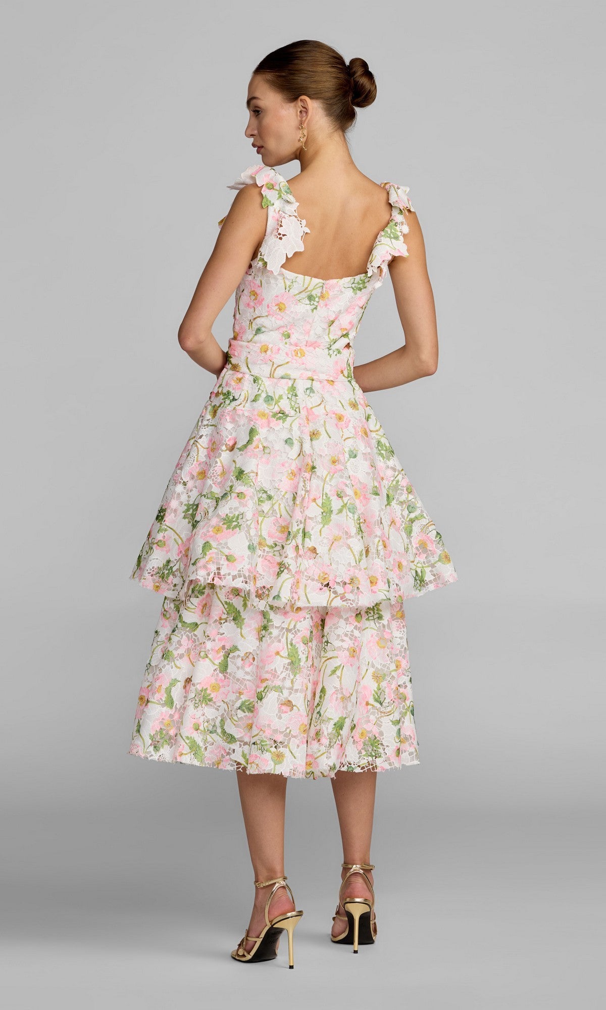 A woman faces away, wearing the Mac Duggal 50112 Midi Party Dress—a sleeveless, tiered dress with a pink floral pattern and ruffled straps—styled as the perfect party look with gold high-heeled sandals, set against a plain gray background.