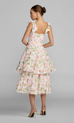 A woman faces away, wearing the Mac Duggal 50112 Midi Party Dress—a sleeveless, tiered dress with a pink floral pattern and ruffled straps—styled as the perfect party look with gold high-heeled sandals, set against a plain gray background.