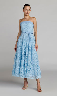 A woman wearing the Mac Duggal 50114 Tea-Length Party Dress in powder blue, featuring floral embroidery lace, a strapless neckline, belted waist, and mid-calf length, stands against a plain gray background.