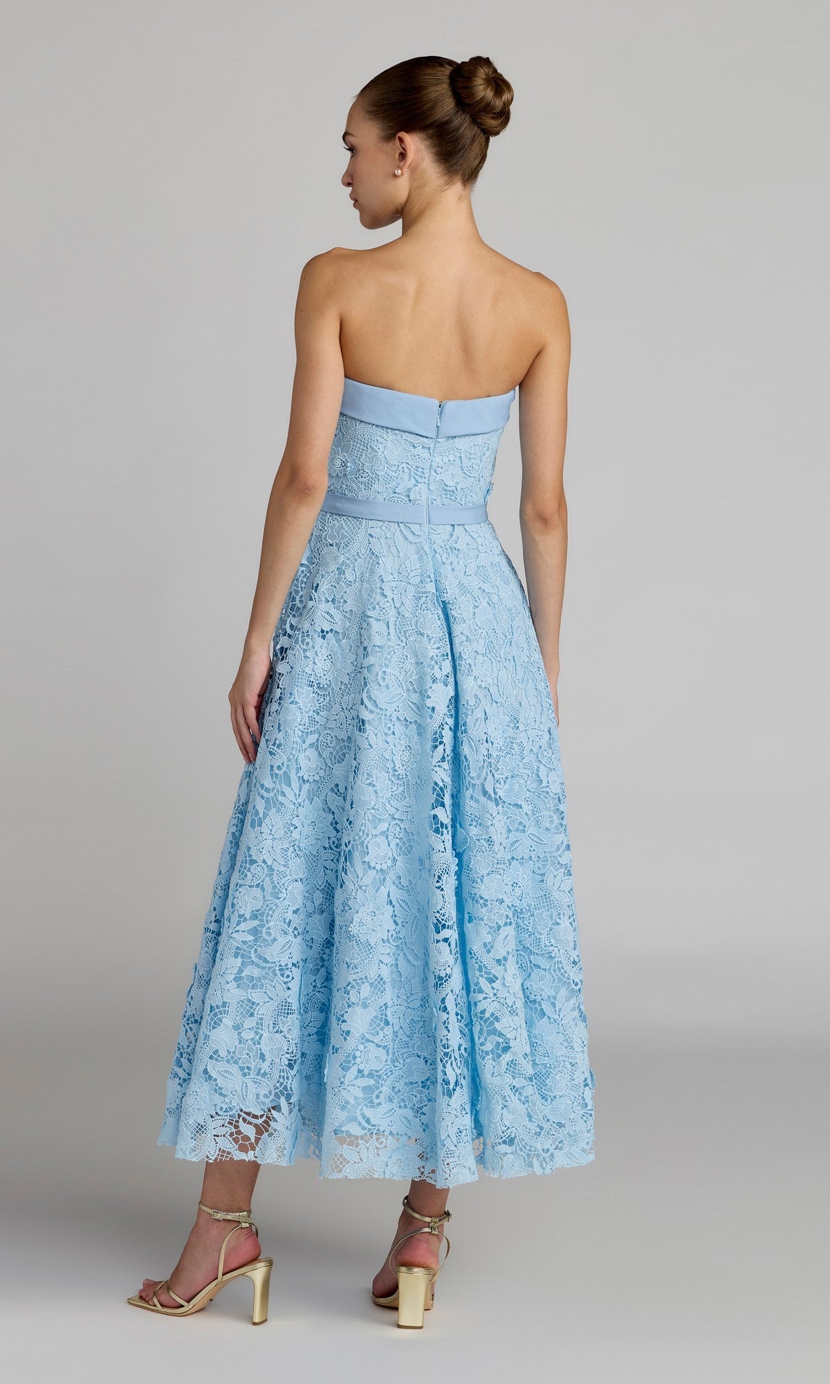 A woman stands facing away from the camera against a plain background, wearing the Mac Duggal 50114 tea-length party dress, which features light blue floral embroidery lace, a strapless neckline, and a flared midi skirt. She completes the look with beige heeled sandals.