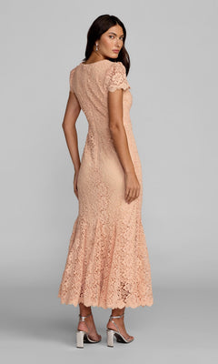 A woman stands facing away against a plain gray background, wearing the Mac Duggal 50116 Tea-Length Party Dress in peach lace with short sleeves, paired with silver high-heeled sandals.