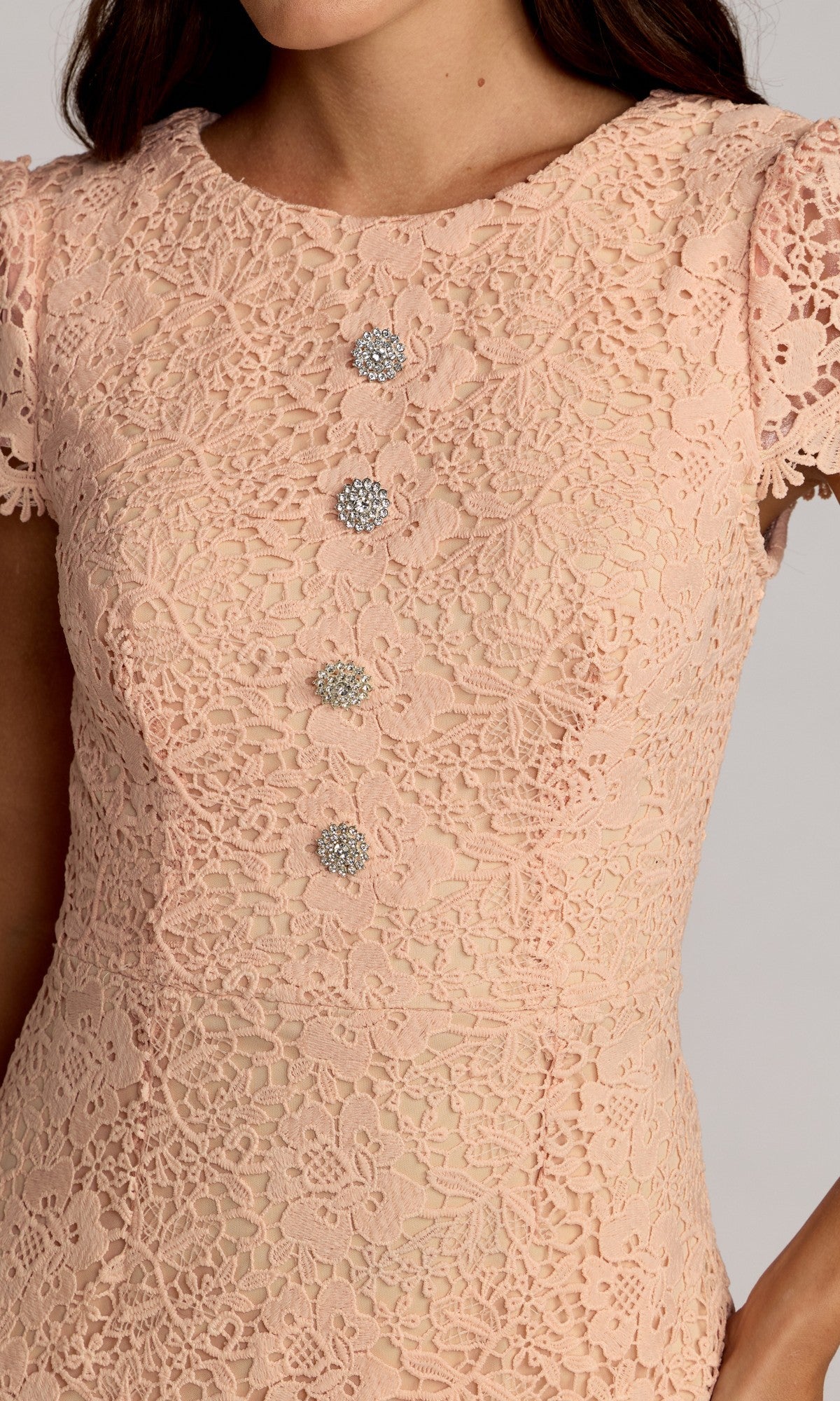 A woman models the Mac Duggal 50116 tea-length party dress in pale pink lace, featuring short sleeves and five decorative silver buttons down the front. Only her upper torso is visible.
