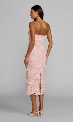 A woman stands facing away, wearing the Mac Duggal 50117 Midi Party Dress in pale pink, featuring a strapless design adorned with delicate 3D floral appliqué and lace details. She pairs it with matching heels and gold earrings against a plain light gray background.