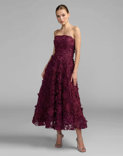 A woman wearing the Mac Duggal 50118 long formal dress in plum, featuring elegant embroidered lace details and a strapless midi silhouette, stands in silver heels against a plain gray background.