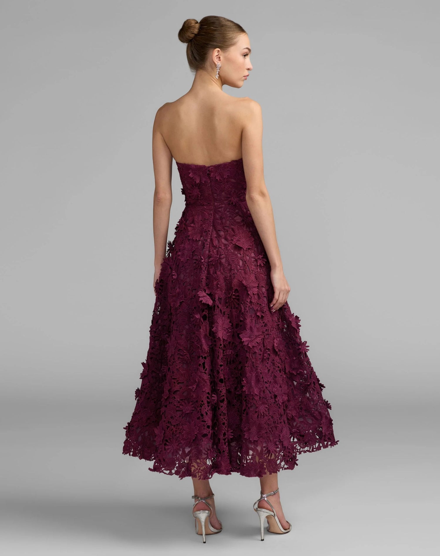A woman wearing the Long Formal Dress: Mac Duggal 50118—a strapless, burgundy embroidered lace tea-length gown with a flared skirt—stands in high heels facing away from the camera against a plain gray background.