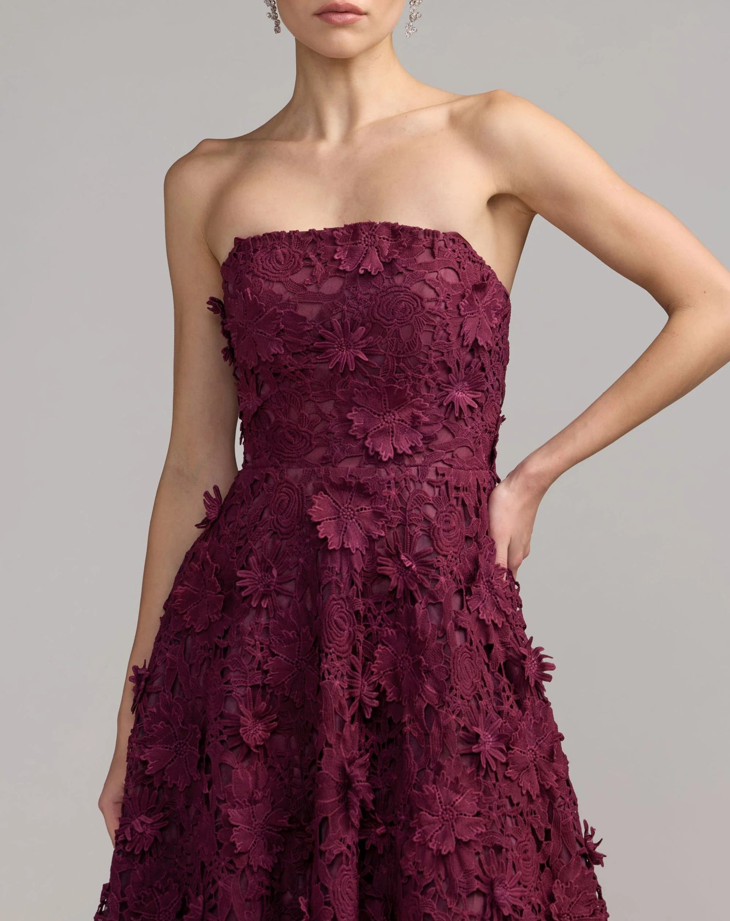 A woman poses with one hand on her hip against a plain background, wearing the Mac Duggal 50118—a long formal dress in rich burgundy, featuring a strapless neckline and delicate embroidered lace detailing.