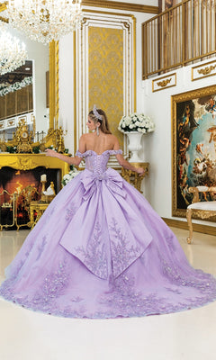 A woman in the Lilac Quinceanera Dress 5011 by Dancing Queen, featuring a ball gown skirt and large bow, stands in an ornate room with gold decor, a chandelier, and a fireplace.