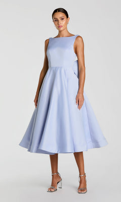 A woman wearing the Tea-Length Party Dress: Mac Duggal 50127 in periwinkle, a sleeveless, knee-length A-line design, paired with silver high-heeled sandals, stands against a plain light background.