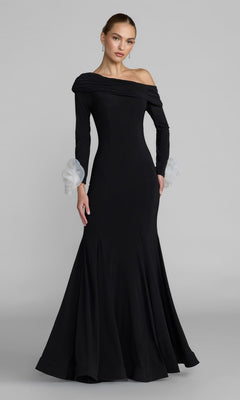 A woman wearing the Long Formal Dress: Mac Duggal 50134 in Black Ivory, featuring a long, fitted mermaid silhouette with an off-the-shoulder neckline, flared hem, and sheer white ruffled cuffs, stands against a plain gray background.