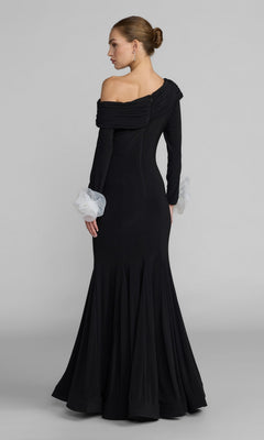 A woman stands facing away against a plain gray background, her hair styled in a neat bun, wearing the Mac Duggal 50134 long formal dress—a black one-shoulder gown featuring sheer white ruffle cuffs.
