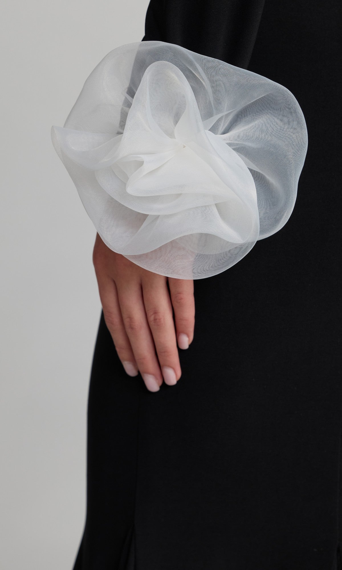 A person wearing the Mac Duggal 50134 long formal dress, a black one-shoulder gown featuring a large sheer white floral detail on the sleeve, standing with their hand at their side.