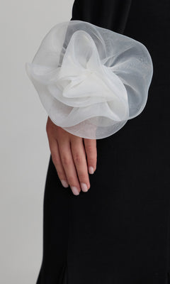 A person wearing the Mac Duggal 50134 long formal dress, a black one-shoulder gown featuring a large sheer white floral detail on the sleeve, standing with their hand at their side.