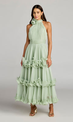 A woman stands against a plain gray background wearing the Mac Duggal 50145 long formal dress in jade, featuring a sleeveless design with a high halter neckline and ruffled midi skirt, paired with gold high-heeled sandals.