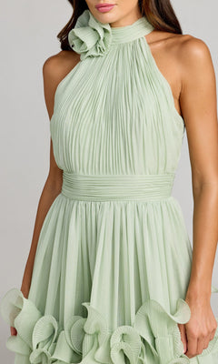 A woman wearing the Long Formal Dress: Mac Duggal 50145, a light green chiffon tea-length gown with a high halter neckline, showcasing pleated fabric and ruffled flower accents at the neck and skirt in Mac Duggal’s signature style.