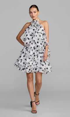 A woman models the Mac Duggal 50171 Short Party Dress—a sleeveless white halter dress with large black polka dots, cascading ruffles, and a floral accent at the neckline—paired with black strappy heels, set against a plain light gray background.