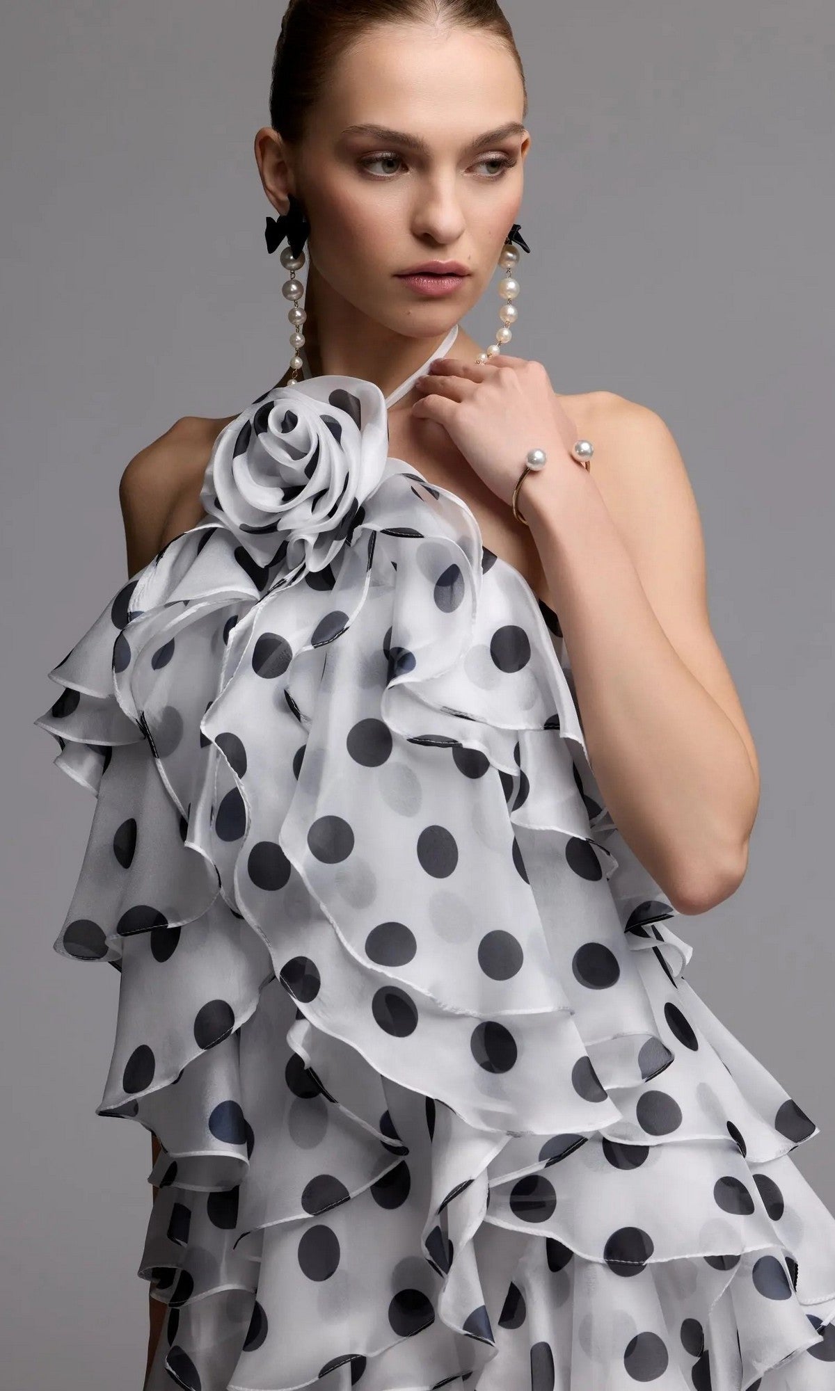 Styled in the Mac Duggal 50171 Short Party Dress—a striking, one-shoulder design featuring black polka dots, dramatic cascading ruffles, and a statement fabric flower—this woman accessorizes with pearl earrings and a bracelet. The plain background highlights the dress’s movement and elegance, creating an effortlessly chic party look.