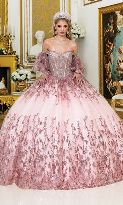 A woman wearing the Quinceanera Dress 5019 by Dancing Queen, featuring a jeweled tiara and ornate floral details with a sheer corset bodice, stands in an opulent gold-decorated room.
