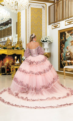 A woman in the Quinceanera Dress 5019 By Dancing Queen, featuring ornate pink fabric and floral details, stands with her back to the camera in an elegant gold-accented room with classical decor and a glowing fireplace.