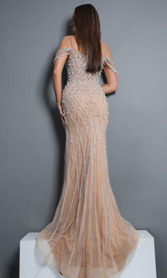 A woman stands on a white platform, facing away, wearing the Jovani 50202 long formal dress—a fitted, nude-colored gown featuring beaded geometric patterns and off-the-shoulder straps—against a plain gray background.