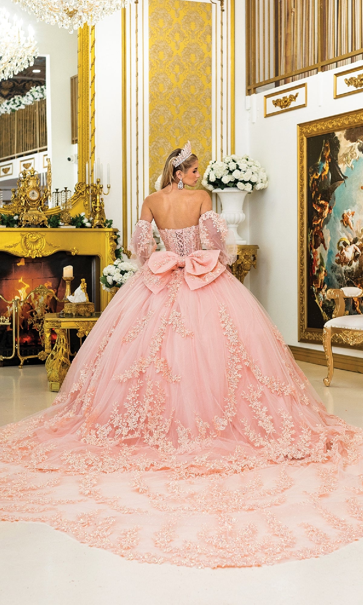 A woman in the Quinceanera Dress 5020 By Dancing Queen, featuring a blush pink corset bodice, stands with her back to the camera in a lavish gold room with a grand fireplace and classical painting.