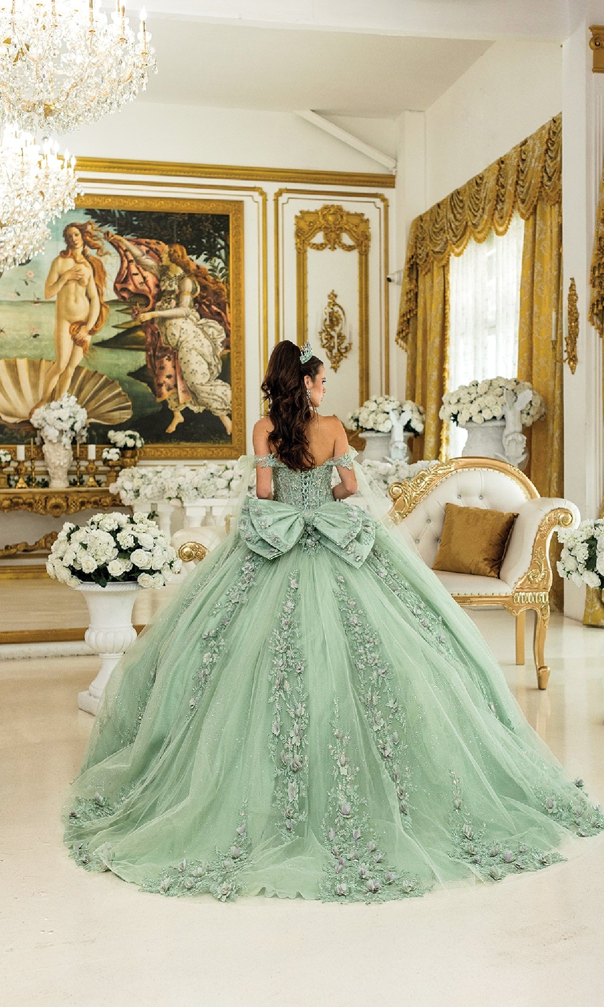 A woman in the Green Quinceanera Dress 5021 by Dancing Queen stands in an ornate room with gold accents, white flower arrangements, and a classical painting on the wall.