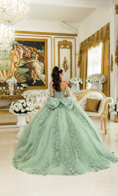 A woman in the Green Quinceanera Dress 5021 by Dancing Queen stands in an ornate room with gold accents, white flower arrangements, and a classical painting on the wall.