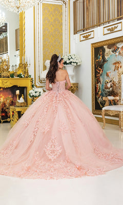 A young woman wears the Quinceanera Dress 5027 By Dancing Queen, featuring a blush pink ball gown and sparkling corset bodice, as she stands with her back to the camera in an ornate gold-accented room.