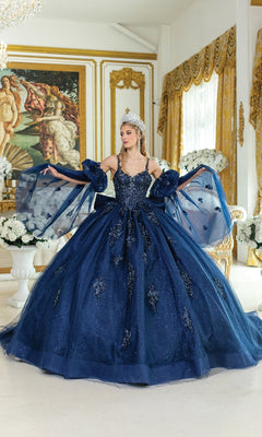 A woman in the Blue Quinceanera Dress 5037 By Dancing Queen and a tiara poses indoors, surrounded by white flowers and ornate gold-trimmed decor with classical paintings in the background.