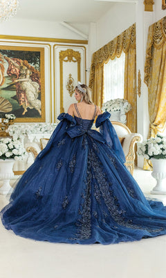A woman in the Blue Quinceanera Dress 5037 By Dancing Queen and a tiara stands in an ornate, gold-accented room with white flowers, facing away from the camera.