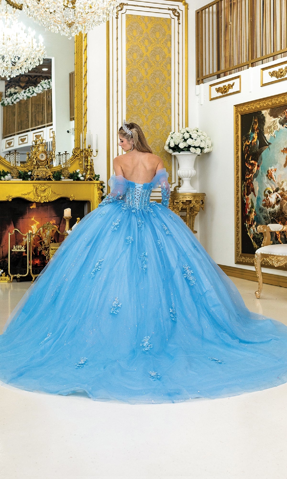 A woman wearing the Blue Quinceanera Dress 5038 by Dancing Queen and a tiara stands with her back to the camera in an ornate, gold-accented room featuring a chandelier, fireplace, and classical décor.