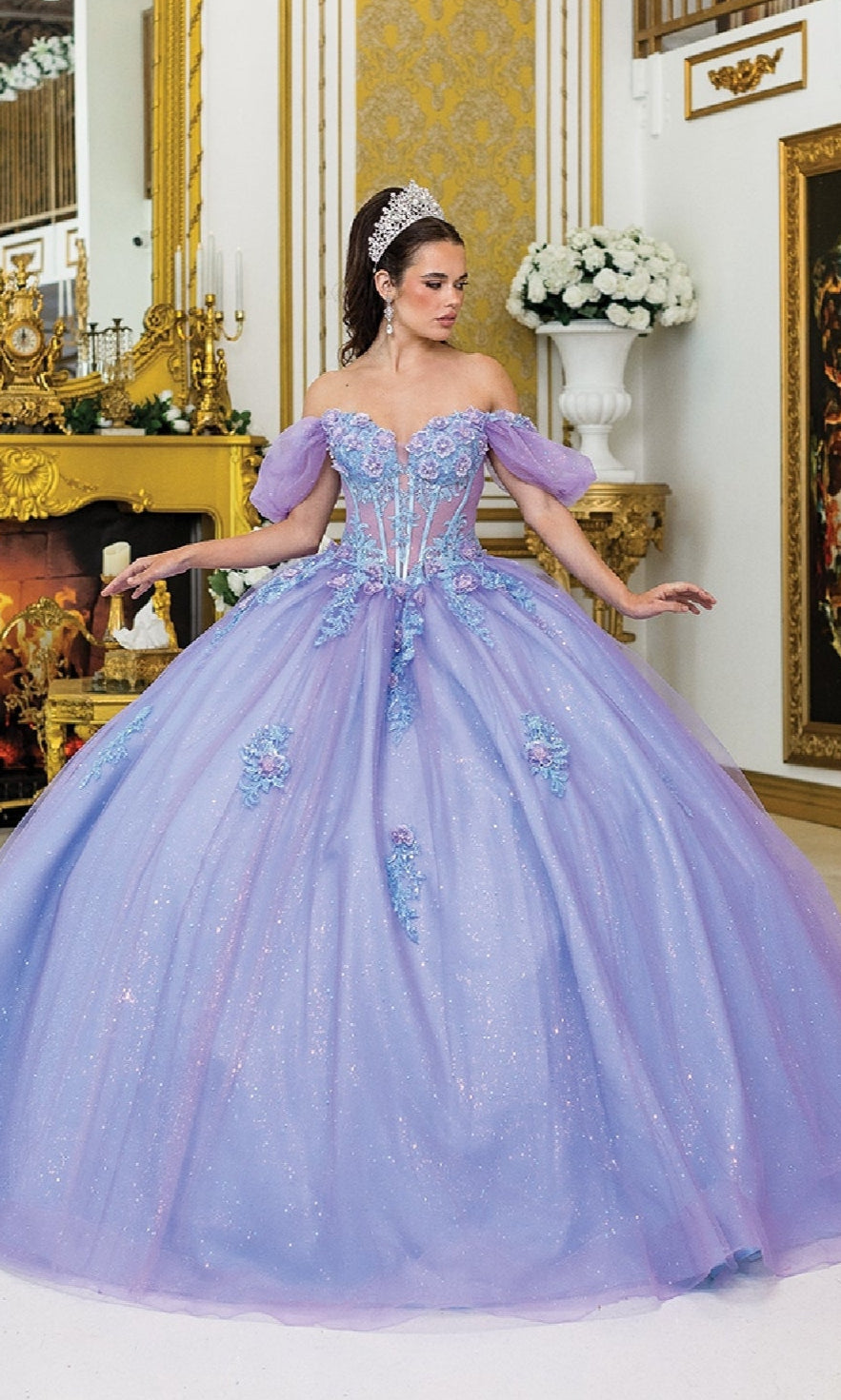 A woman wearing the Quinceanera Dress 5039 By Dancing Queen—a large, off-the-shoulder gown with a glitter tulle skirt and floral details—stands in an ornate, gold-decorated room with a tiara.