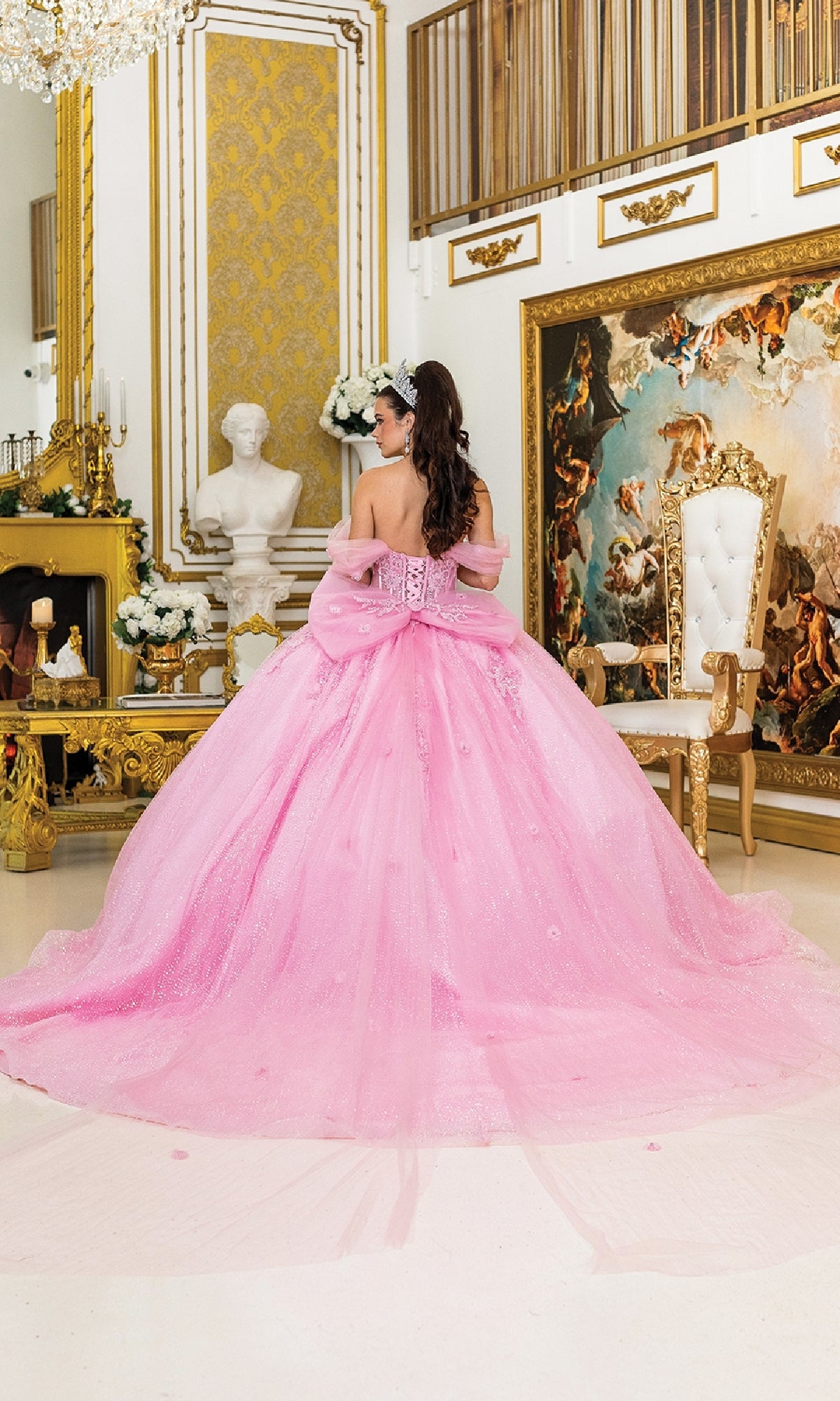 A woman wears the Quinceanera Dress 5040 By Dancing Queen, featuring a dramatic pink ball gown skirt and bow, standing in an ornate room with gold accents, a white chair, and classical artwork.