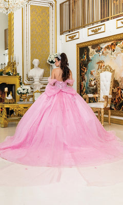 A woman wears the Quinceanera Dress 5040 By Dancing Queen, featuring a dramatic pink ball gown skirt and bow, standing in an ornate room with gold accents, a white chair, and classical artwork.
