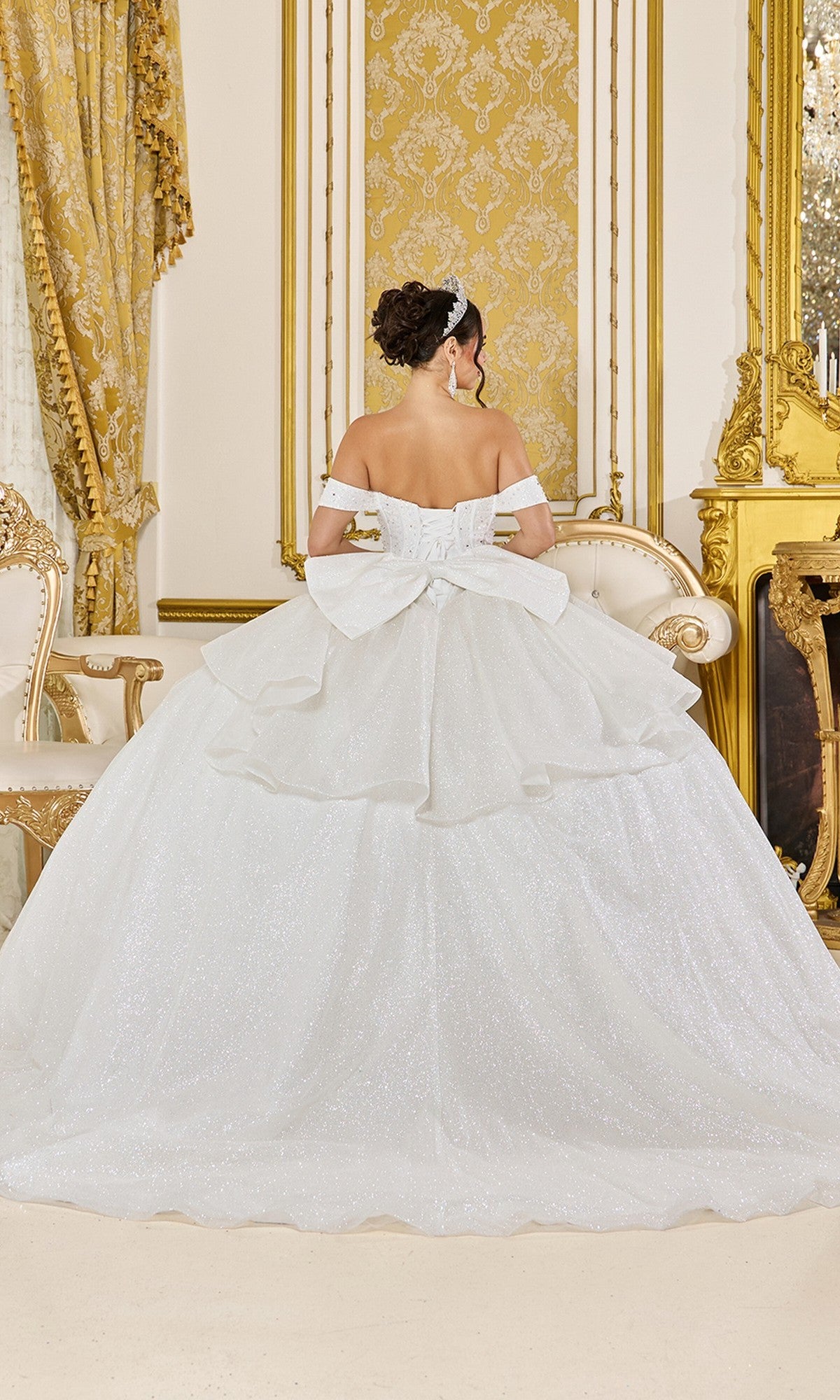 A woman stands facing away in an ornate, gold-accented room with elegant furniture and drapery, wearing the Quinceanera Dress 5043 By Dancing Queen—a white off-the-shoulder tulle ballgown featuring a large bow.