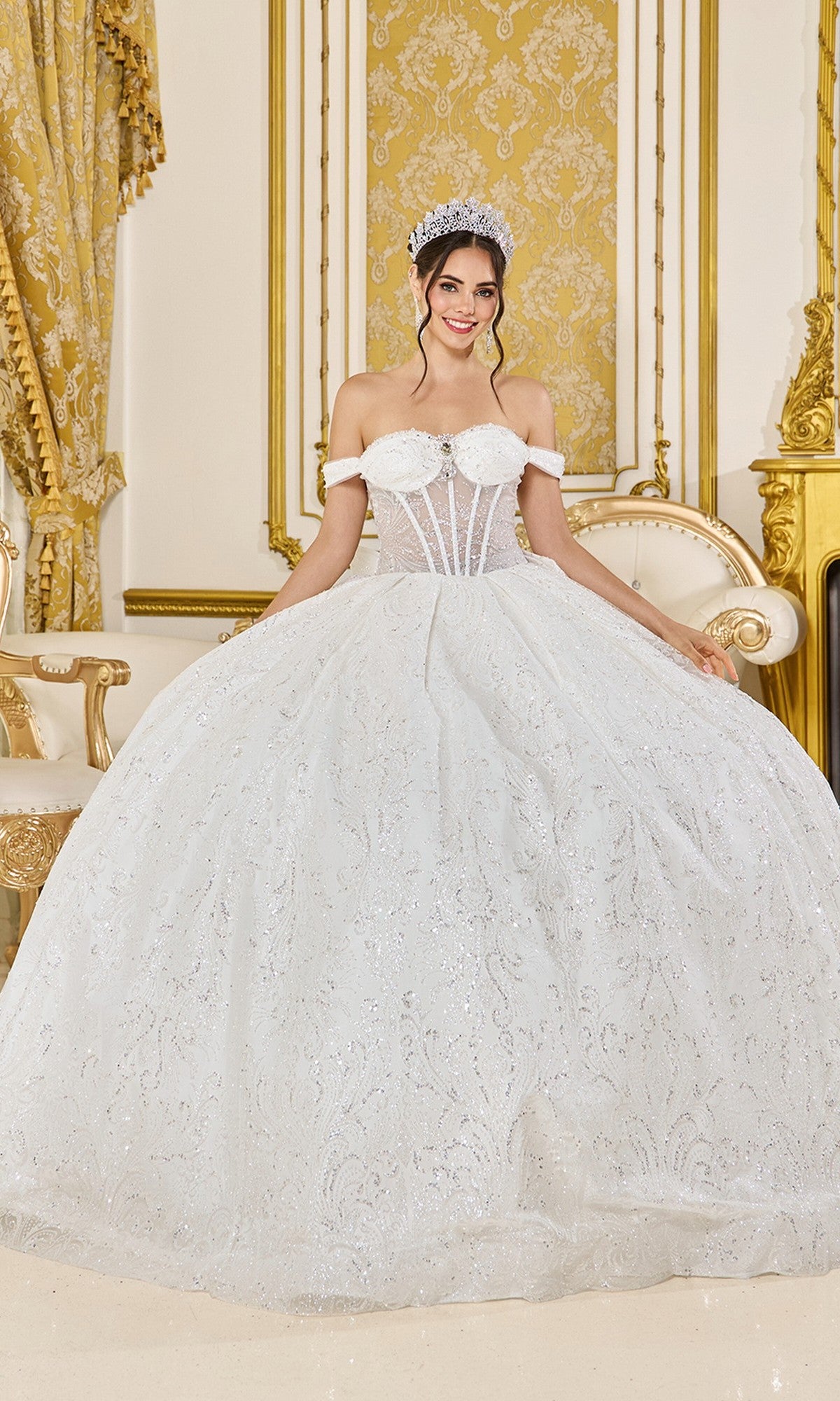 A woman wearing the Quinceanera Dress 5043 by Dancing Queen in Off White, featuring a glittering tulle ballgown and corset bodice, stands in an ornate room with gold accents and patterned walls, smiling at the camera.