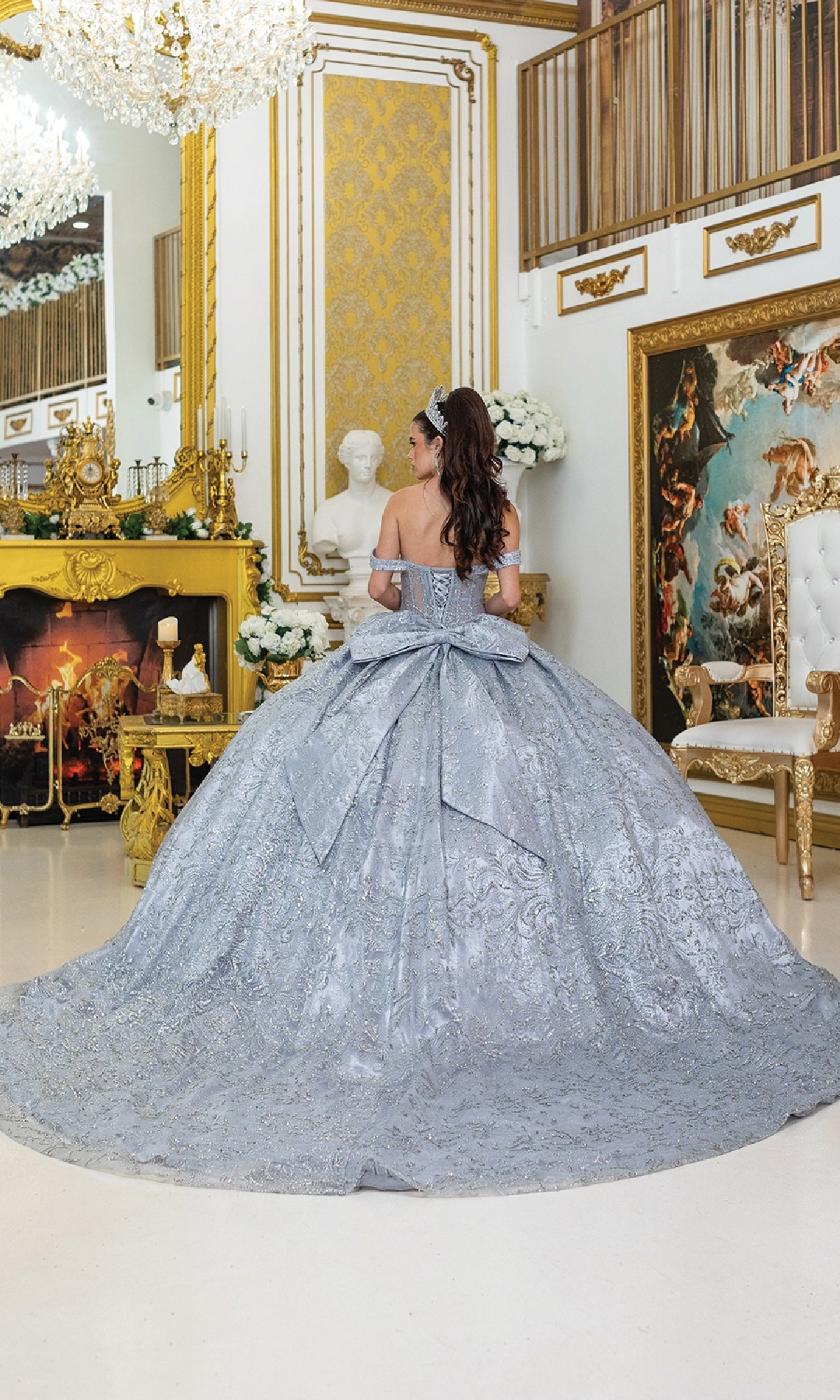 A woman in the Quinceanera Dress 5043 by Dancing Queen—a silver tulle ballgown featuring a corset bodice and a large bow—stands in an ornate room adorned with gold decor, a fireplace, and classical artwork.