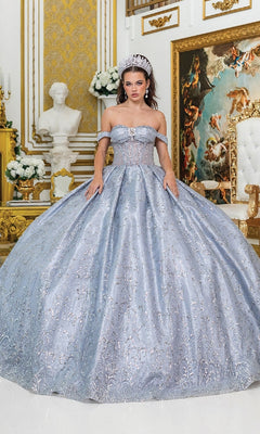 A woman wearing the Quinceanera Dress 5043 by Dancing Queen—a blue tulle ballgown with a corset bodice and a tiara—stands in an ornate room adorned with gold accents, white flowers, a white statue, and a large painting in the background.