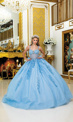 A person in the Quinceanera Dress 5046 By Dancing Queen, featuring a blue ball gown and corset bodice with tiara, stands in an ornate gold-accented room with a fireplace, chandeliers, and classical decor.