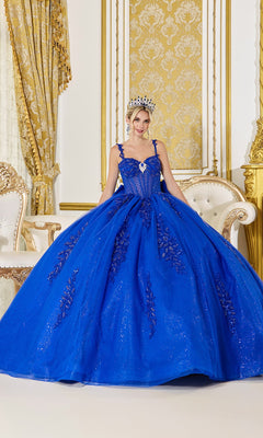 A young woman stands indoors, surrounded by ornate gold and white furniture and decor, wearing the Quinceanera Dress 5046 By Dancing Queen in royal blue. The gown features a glitter tulle skirt and an elegant corset bodice, while her tiara sparkles softly in the light.
