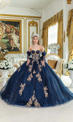 A woman wearing the Quinceanera Dress 5051 By Dancing Queen—a navy blue and gold embellished glitter tulle ball gown with a tiara—stands in an ornate room with gold accents, white flowers, and a grand classical painting.